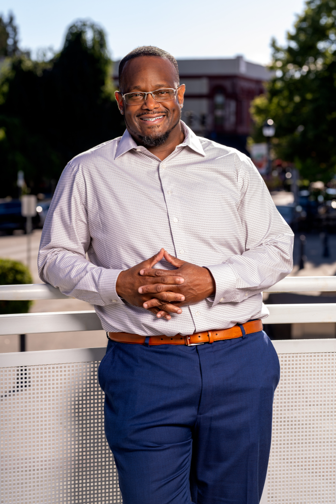A man wearing glasses, a light-colored dress shirt, and blue pants stands outdoors, smiling and leaning against a white railing with trees and buildings blurred in the background.