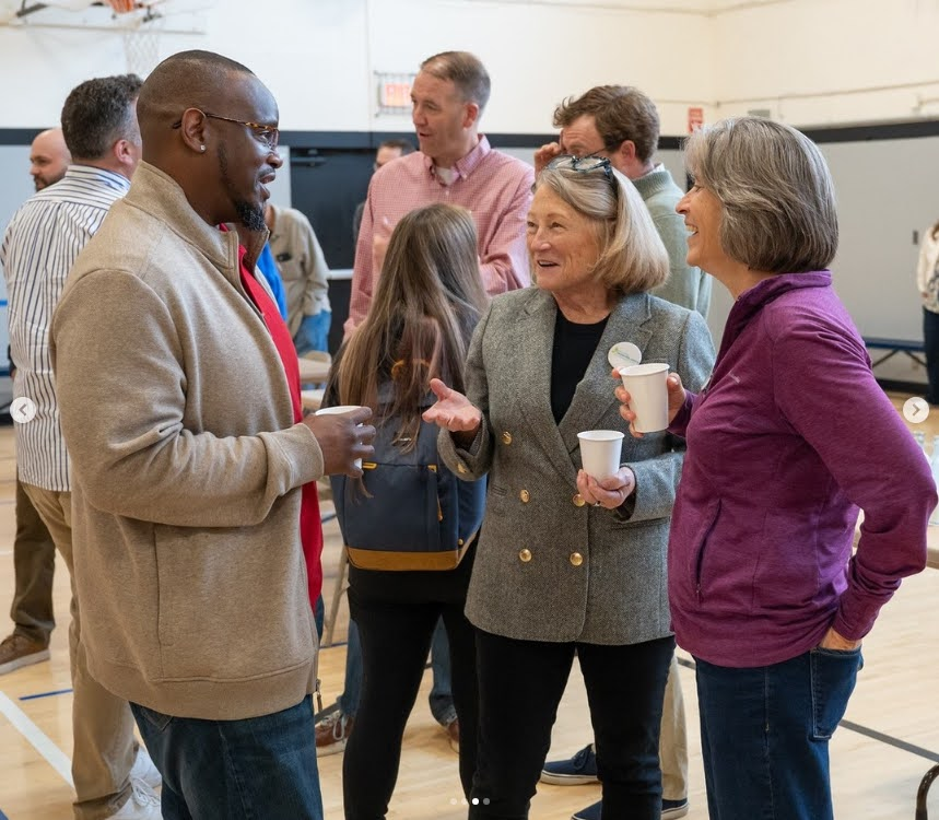 A group of adults stand in a gymnasium, chatting and holding cups. Three people in the foreground are smiling and engaged in conversation, suggesting a friendly, casual event. Other people are mingling in the background.