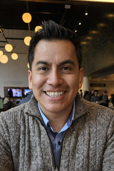 A man with dark hair and a friendly smile sits indoors, wearing a brown zip-up sweater over a blue collared shirt. Warm, round lights hang from the ceiling, and people are visible in the background.