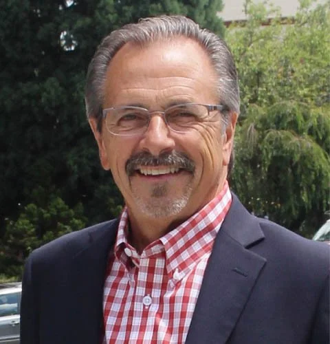 A smiling middle-aged man with glasses, gray hair, and a mustache wears a navy blazer over a red and white checkered shirt, standing outdoors with trees in the background.