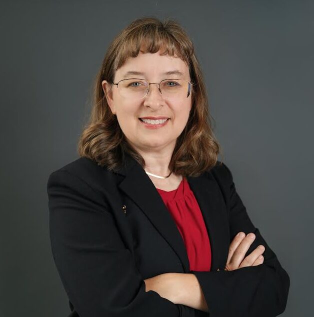 A woman with shoulder-length brown hair, glasses, and a red blouse under a black blazer stands with arms crossed, smiling, in front of a plain gray background.