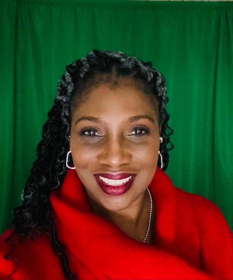A woman with dark curly hair and streaks of gray smiles, wearing a bright red top and hoop earrings, standing in front of a green background.