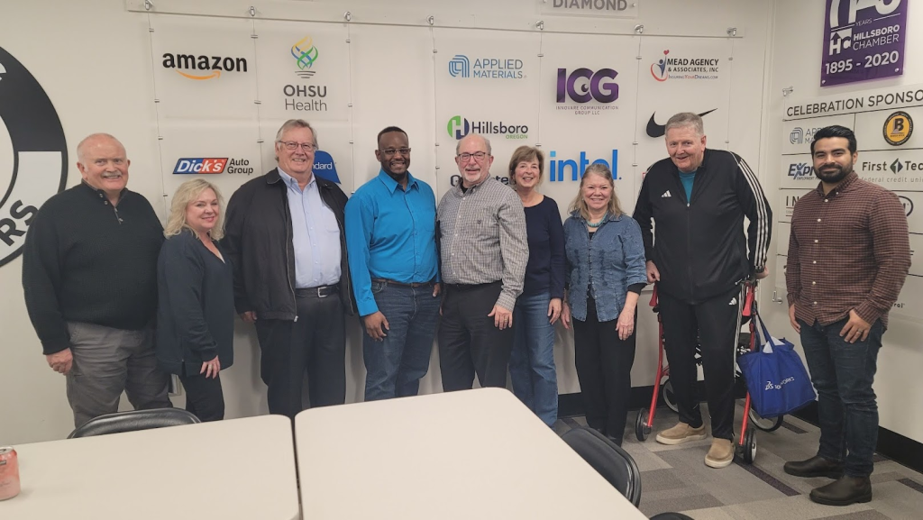 Ten people stand smiling in front of a wall displaying various sponsor logos, including Amazon, OHSU Health, and Applied Materials. They are casually dressed and standing in a brightly lit indoor setting.