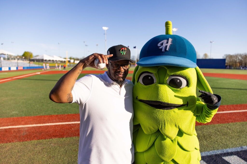 A man in glasses and a white polo stands on a baseball field next to a green mascot wearing a blue hat. Both are smiling and pointing at their hats, with clear skies in the background.