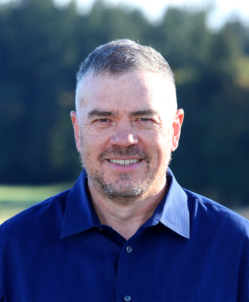 A man with short gray hair and light stubble is smiling outdoors. He is wearing a blue collared shirt, and the background is blurred greenery.