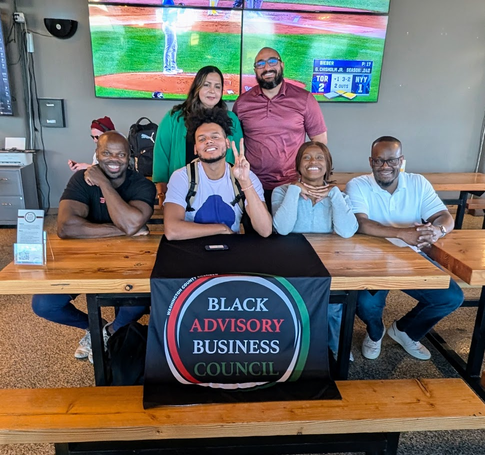 Six people sit and smile at a table with a Black Advisory Business Council banner, posing for a group photo in a modern restaurant. Two large TVs behind them show a baseball game.
