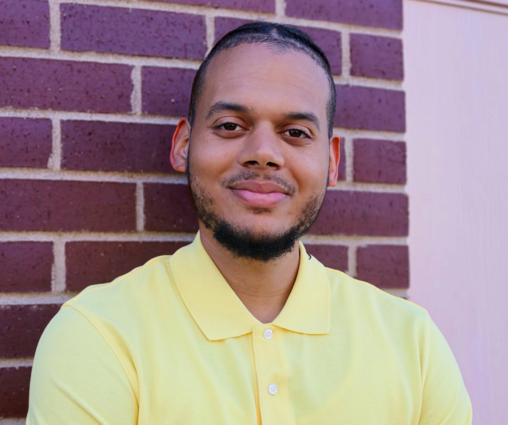 A man in a yellow polo shirt stands in front of a red brick wall, smiling softly at the camera.