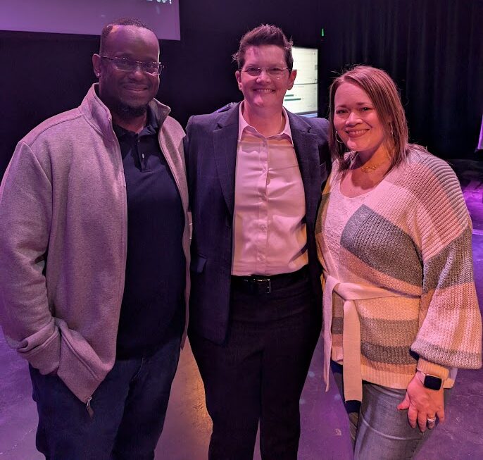 Three adults stand together smiling indoors, with two men and one woman. Behind them, a screen displays a news broadcast with election coverage. The setting appears to be a dimly lit event space or studio.