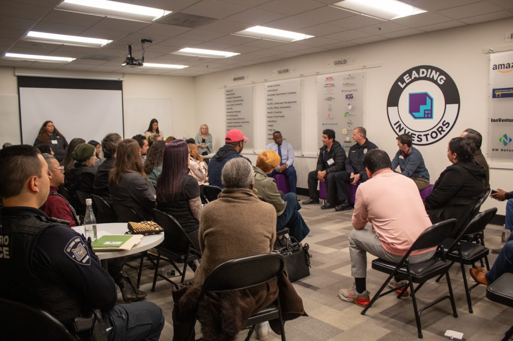 A group of people sit in chairs facing a panel discussion in a conference room. A woman stands near a projector screen, while others listen attentively. Company logos are displayed on the walls.