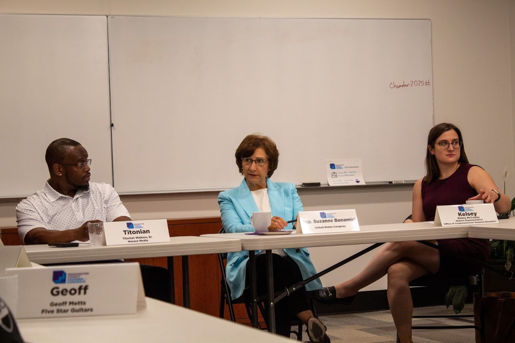 Three people sit at a conference table in discussion. The man on the left wears glasses and a patterned shirt, the woman in the center wears a light blue suit, and the woman on the right wears a burgundy dress. A whiteboard is in the background.
