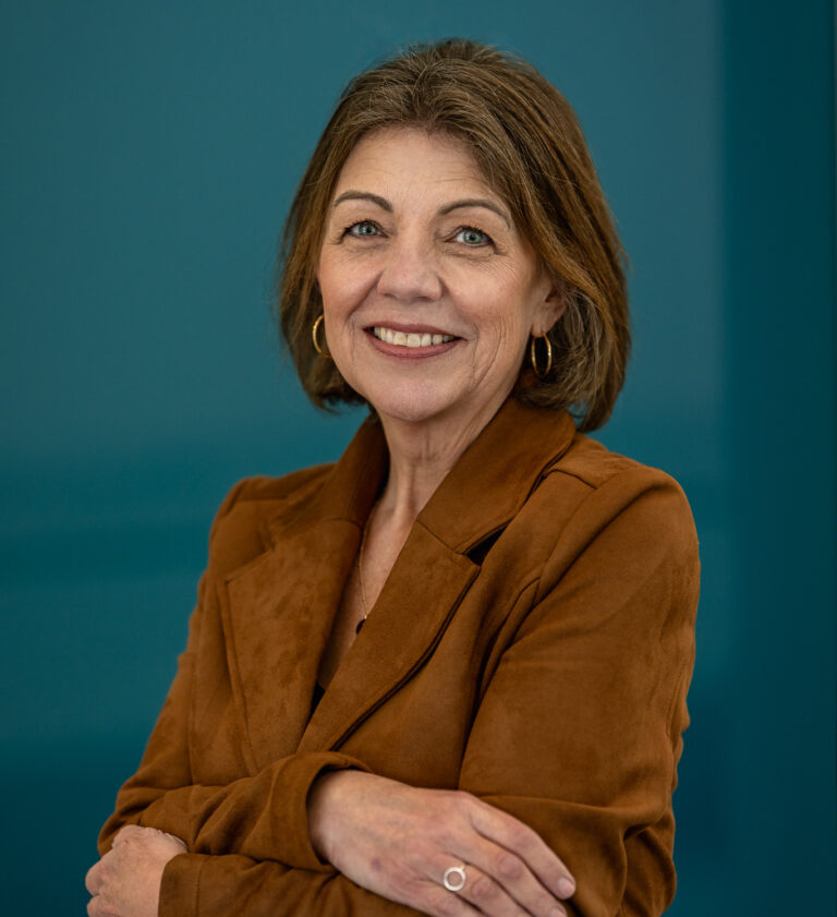 A woman with shoulder-length brown hair, wearing a brown suede jacket and gold hoop earrings, stands smiling with arms crossed against a solid teal background.