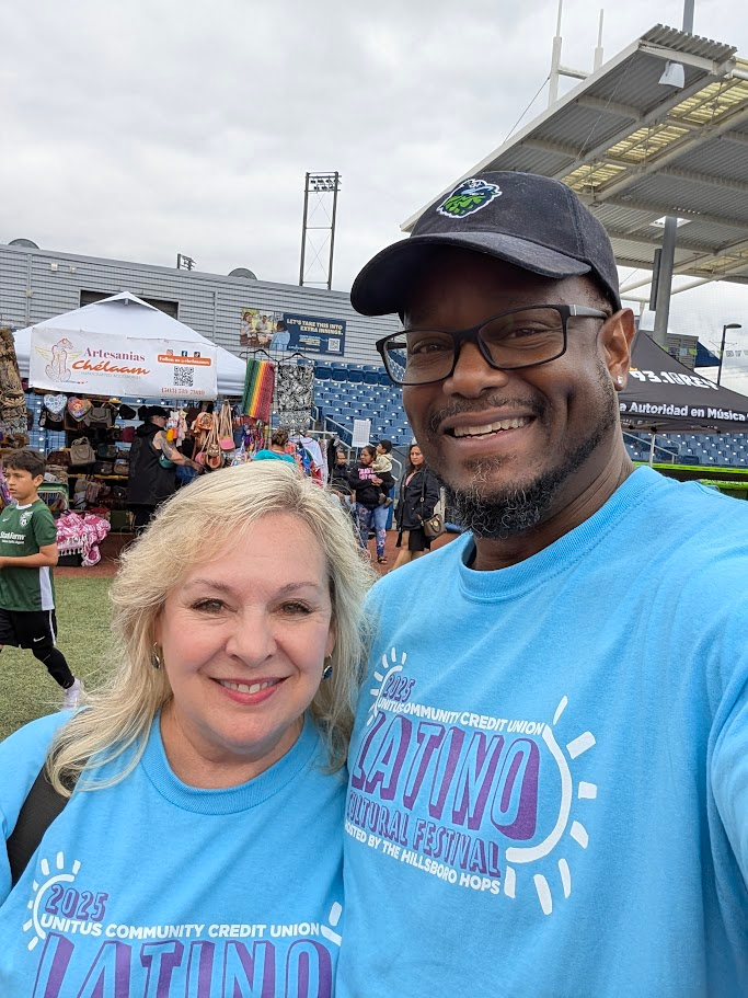Two people wearing blue “Latino Cultural Festival” T-shirts smile at the camera at an outdoor event; behind them are vendor tents, colorful items for sale, and a stadium structure.