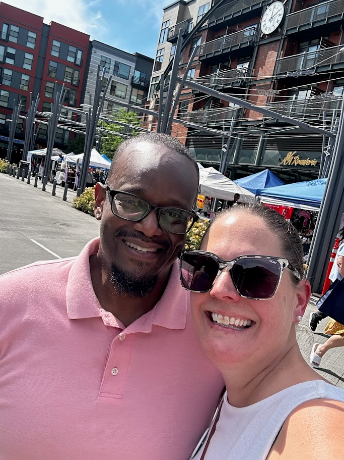 A smiling man and woman pose for a selfie outdoors at a market, with tents, people, and modern buildings in the background on a sunny day.