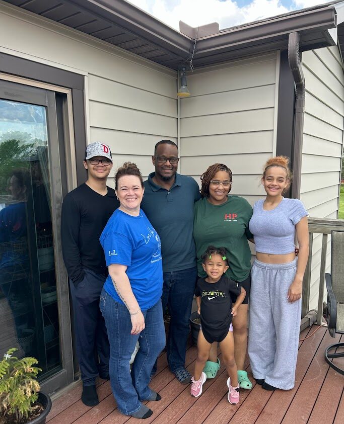 Six people stand together smiling on a wooden deck outside a house. One young child stands in front, while five adults or teens stand behind her. There is a potted plant in the corner and a partly cloudy sky above.