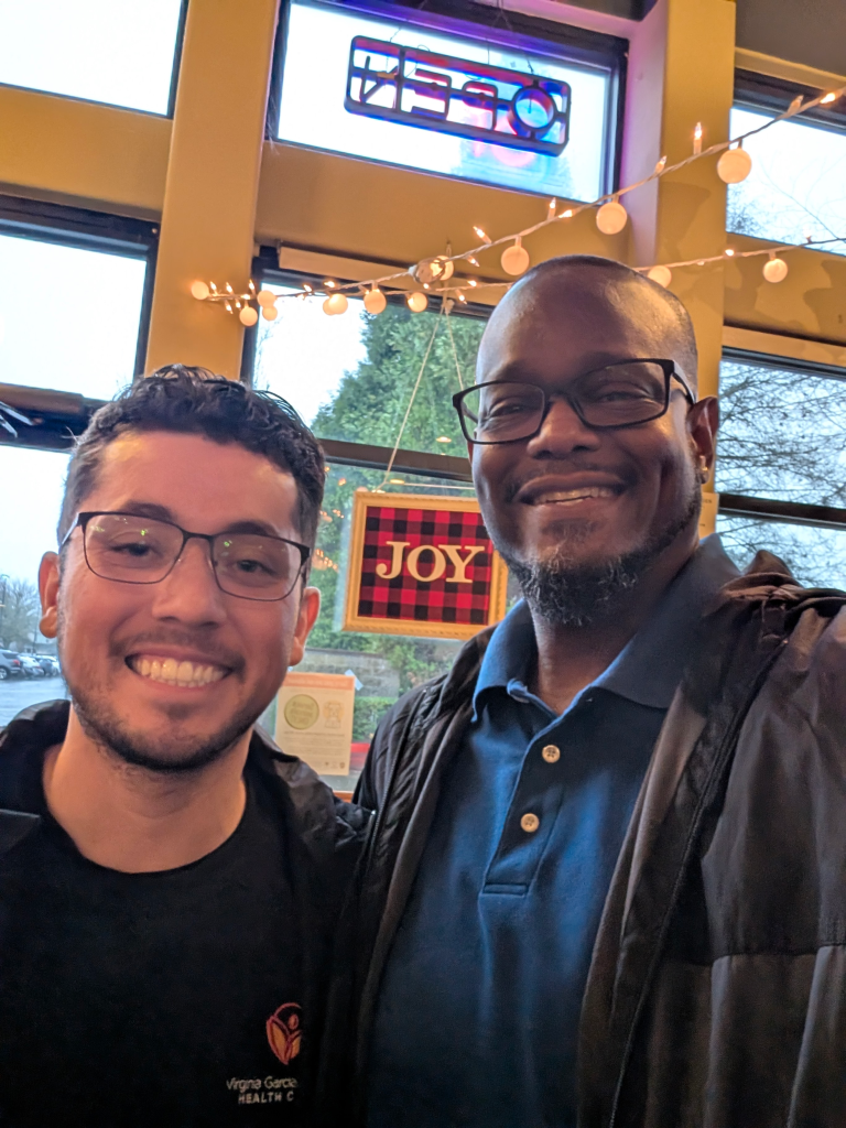 Two men smiling and posing for a selfie inside a cafe. Behind them, a window displays “OPEN” and a festive “JOY” sign with string lights. It appears to be daytime outside.
