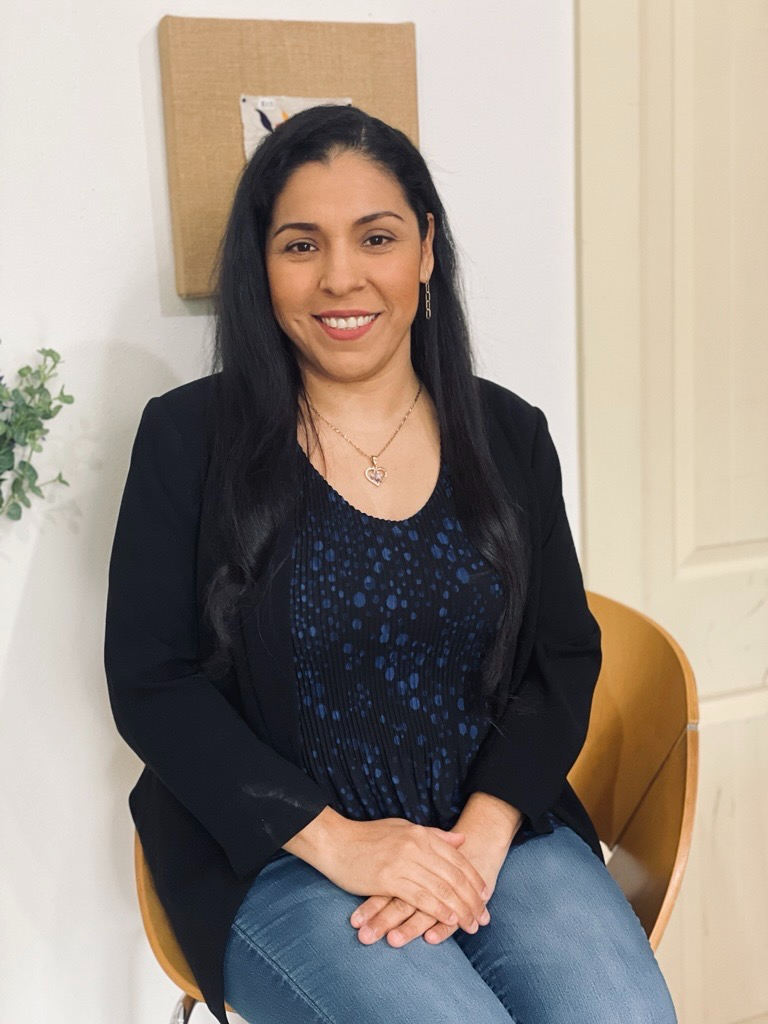 A woman with long dark hair, wearing a black blazer, blue patterned top, and jeans, sits and smiles at the camera. She is seated on a wooden chair in a bright, modern room with neutral decor.