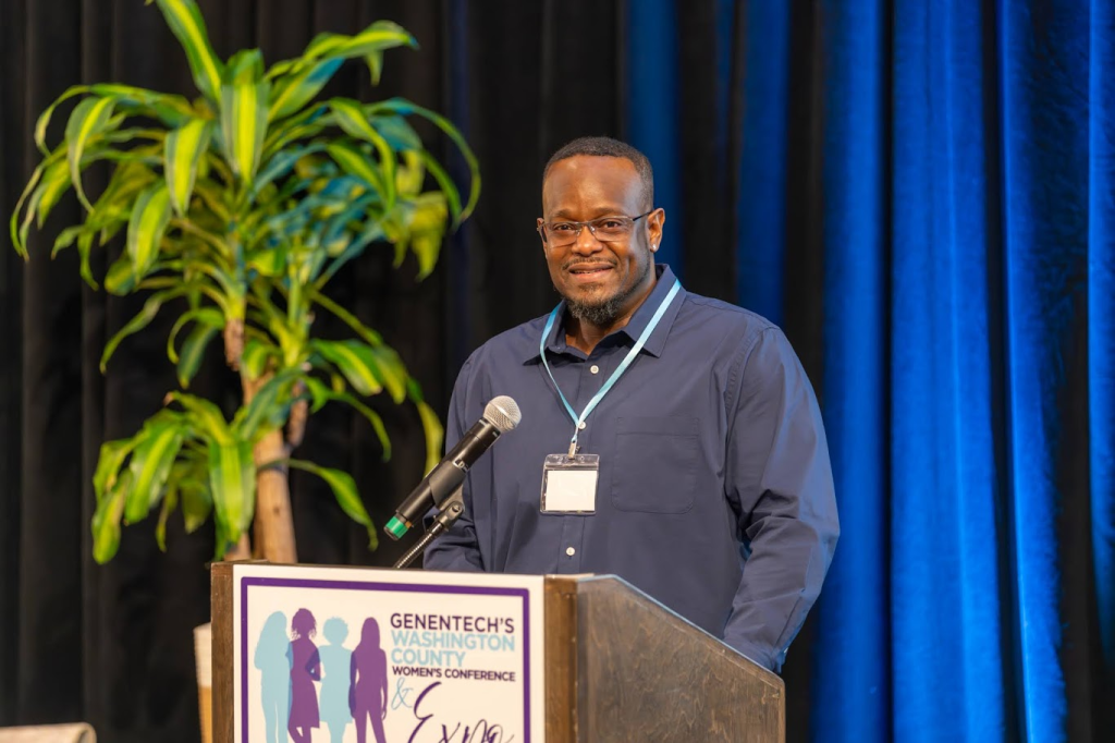 A man wearing glasses and a blue shirt stands at a podium with a microphone, smiling. The podium displays a sign for Genentech's Washington County Women's Conference. A large plant is visible in the background.