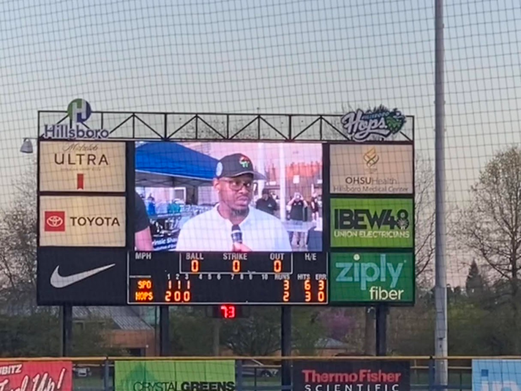 A large stadium scoreboard shows a man in a baseball cap and glasses speaking into a microphone, with game stats for the Hops and SPO teams visible below him. Advertisements and a net are in the background.