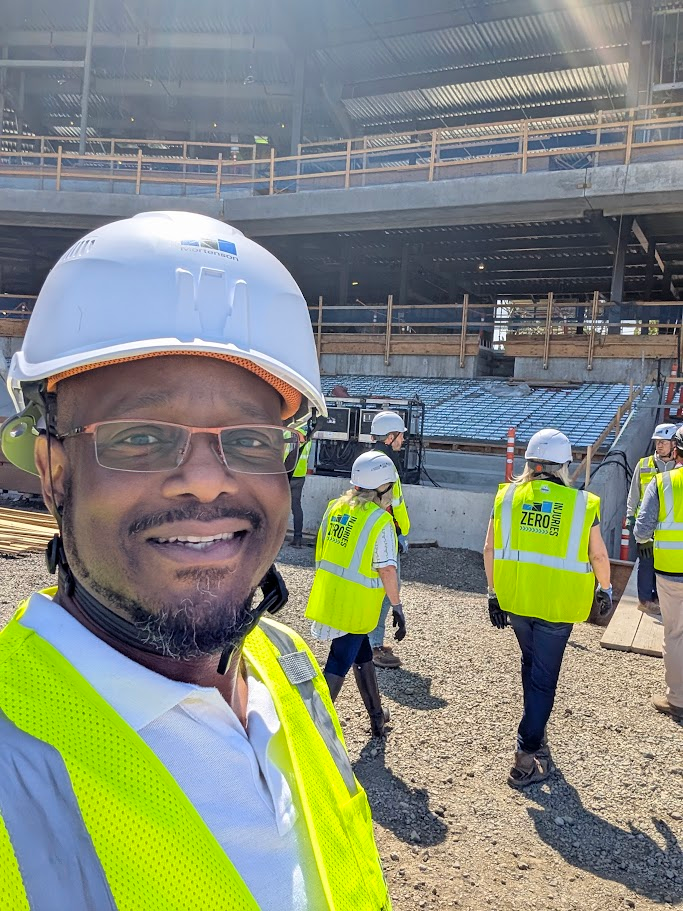 A man wearing a white hard hat, glasses, and a yellow safety vest smiles at the camera on a construction site. Behind him, several workers in similar gear walk toward a partially built structure.