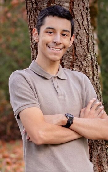 A young man with short dark hair stands outdoors in front of a tree, smiling with his arms crossed. He is wearing a light brown polo shirt and a black wristwatch. The background features greenery and autumn leaves.