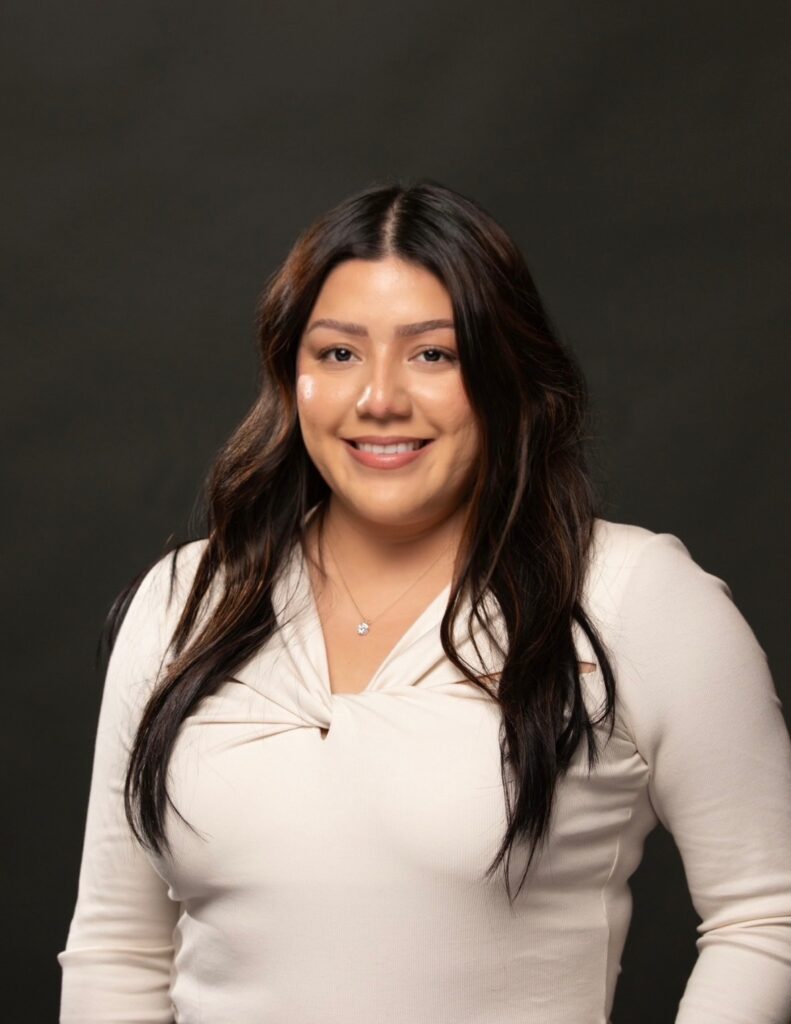 A woman with long, dark hair wearing a cream-colored top smiles in front of a plain dark background. She faces the camera, with her arms relaxed by her sides.