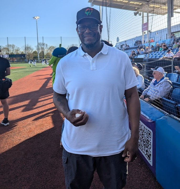 A man in a white polo shirt and black cap stands on a baseball field near the stands, holding a baseball. Spectators sit in blue seats and a game is in progress in the background under a sunny sky.