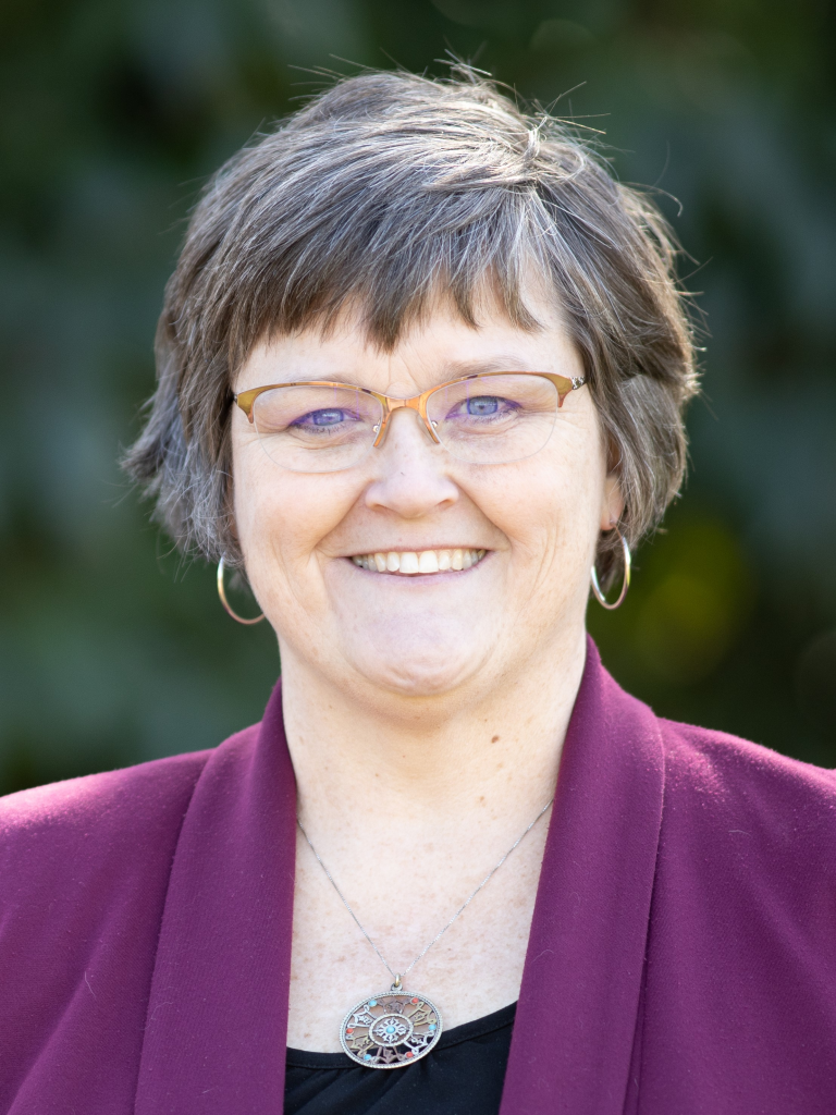 A smiling woman with short, grayish-brown hair, wearing glasses, a maroon blazer, hoop earrings, and a round pendant necklace, standing outdoors with a blurred green background.