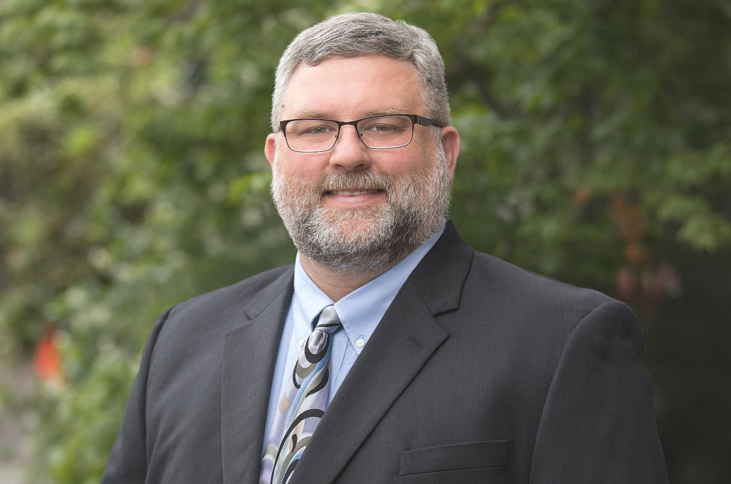 A man with gray hair and beard, wearing glasses, a dark suit, and a patterned tie, stands outdoors in front of green foliage, smiling at the camera.