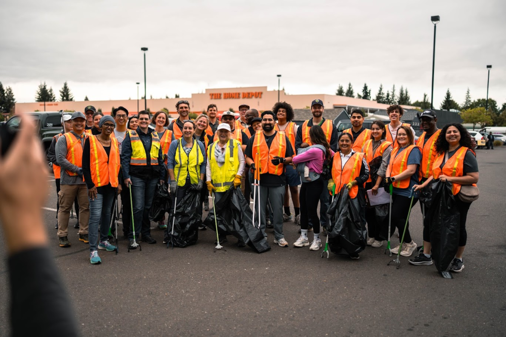 A diverse group of volunteers wearing safety vests poses together with trash bags and litter pickers in a parking lot, with a Home Depot store visible in the background.