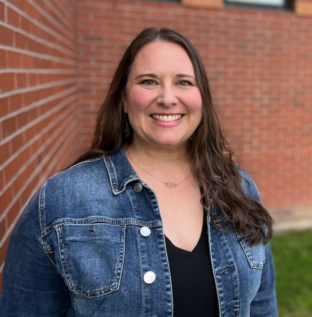 A woman with long brown hair, wearing a denim jacket and black top, smiles while standing outside in front of a brick building with windows and a grassy area.