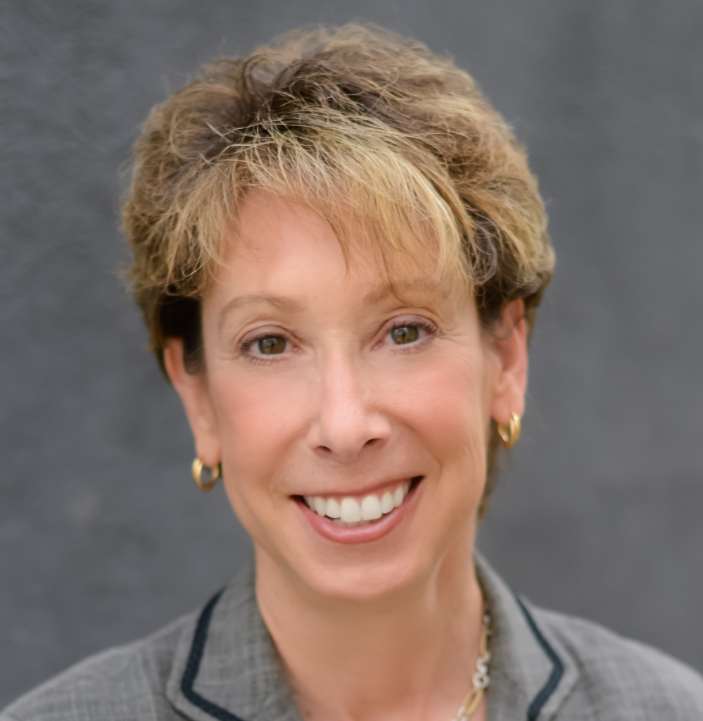 A woman with short, light brown hair and highlights, wearing gold hoop earrings and a gray blazer, smiles at the camera against a plain gray background.