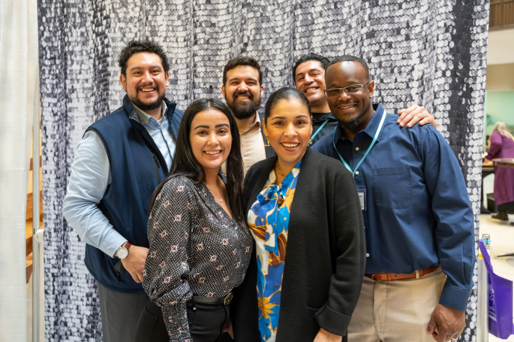 Six adults stand close together, smiling at the camera. They are wearing business casual attire and standing in front of a silver sequin backdrop. The atmosphere appears friendly and cheerful, possibly at a work or social event.
