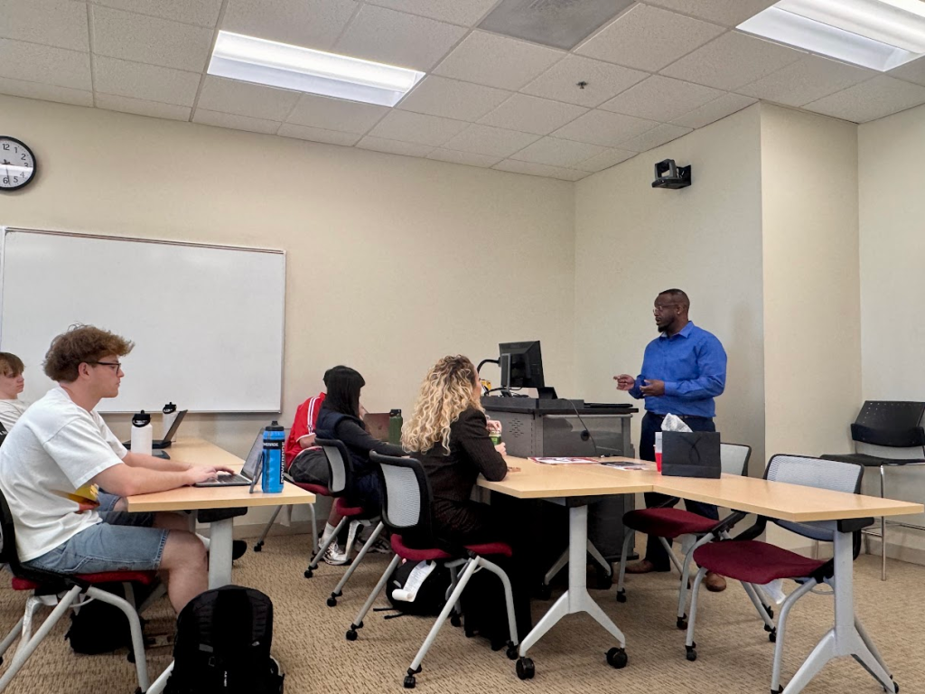 A teacher in a blue shirt stands at a podium addressing four students seated at desks with laptops and notebooks in a classroom with a whiteboard and clock on the wall.