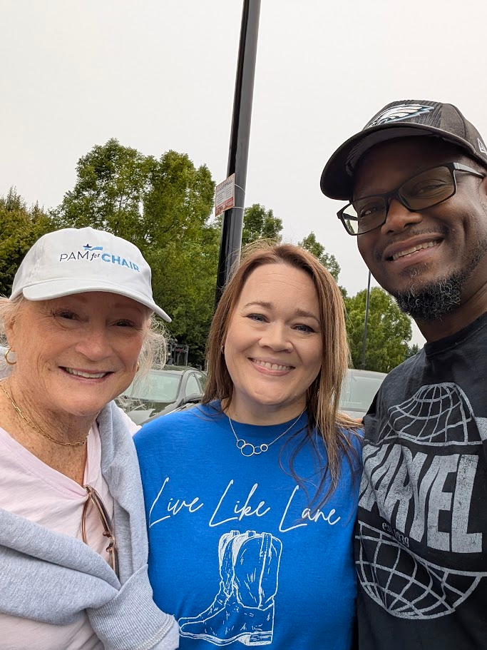 Three people stand close together outdoors, smiling at the camera. The woman on the left wears a white cap and light pink top, the woman in the middle wears a blue shirt, and the man on the right wears glasses and a black T-shirt. Trees and cars are visible in the background.