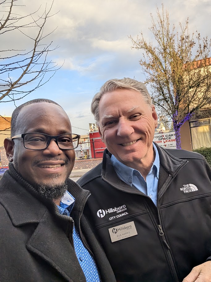 Two men smiling outdoors, both wearing jackets over collared shirts. The man on the right has a "Hillsboro City Council" badge. Trees and buildings are visible in the background under a partly cloudy sky.