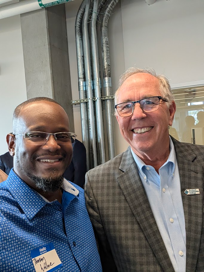 Two men smiling and posing for a photo at an indoor event. One wears glasses and a blue shirt with a name tag, the other has glasses and a gray checked jacket. Exposed pipes and people are visible in the background.