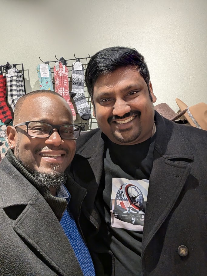 Two men smile for a selfie indoors. Behind them, patterned socks and hats are displayed on a wall rack. Both wear dark jackets; one has glasses and a blue shirt, the other has a T-shirt with a graphic print.