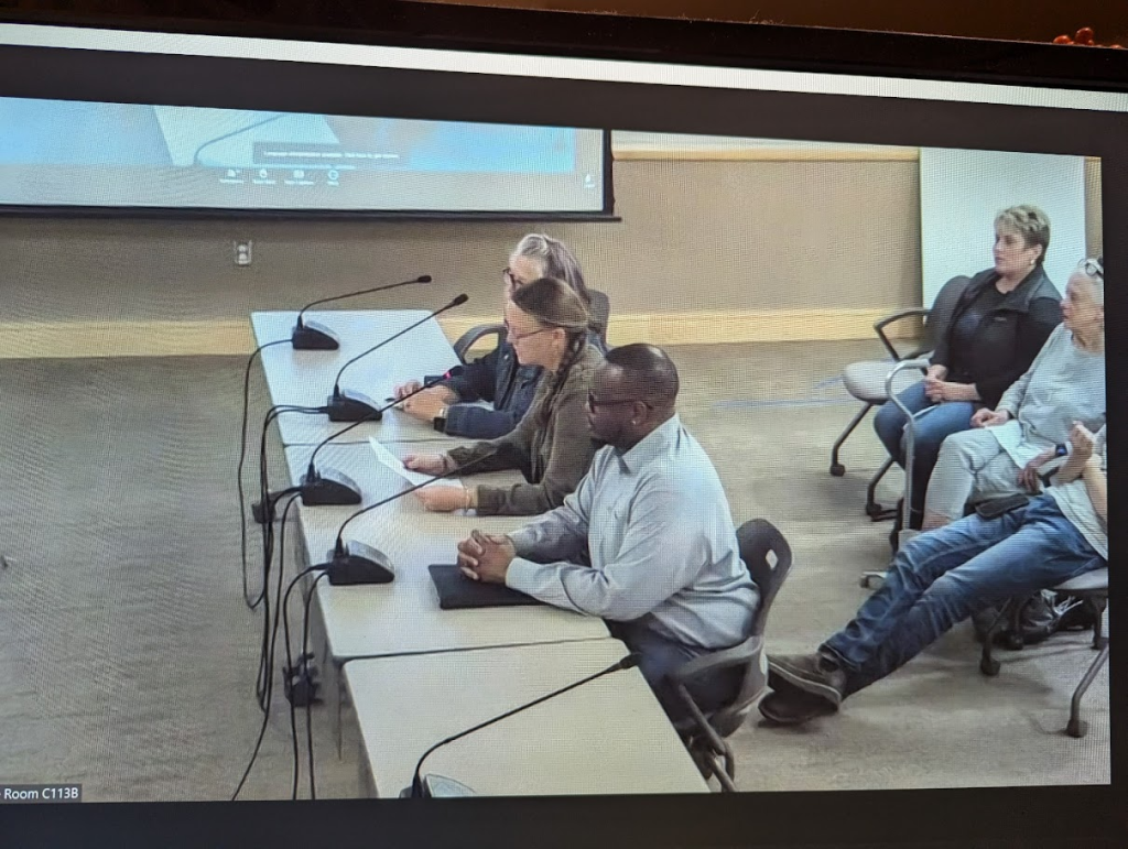 Four people are seated at a conference table with microphones in front of them, speaking to an audience. Three others are seated against the wall in the background, observing the meeting. A projection screen is visible on the wall.
