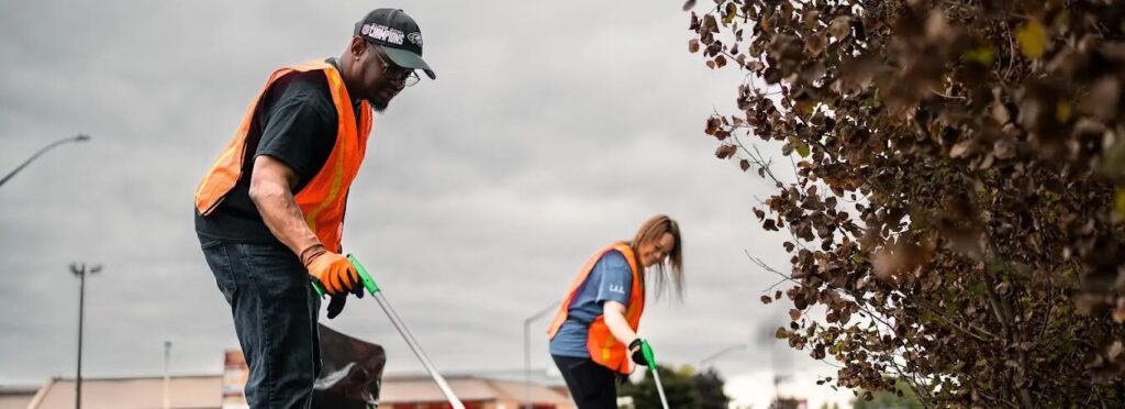 Two people wearing orange safety vests and gloves use grabbers to pick up litter near railroad tracks, with a pile of trash in the foreground and cloudy skies overhead.