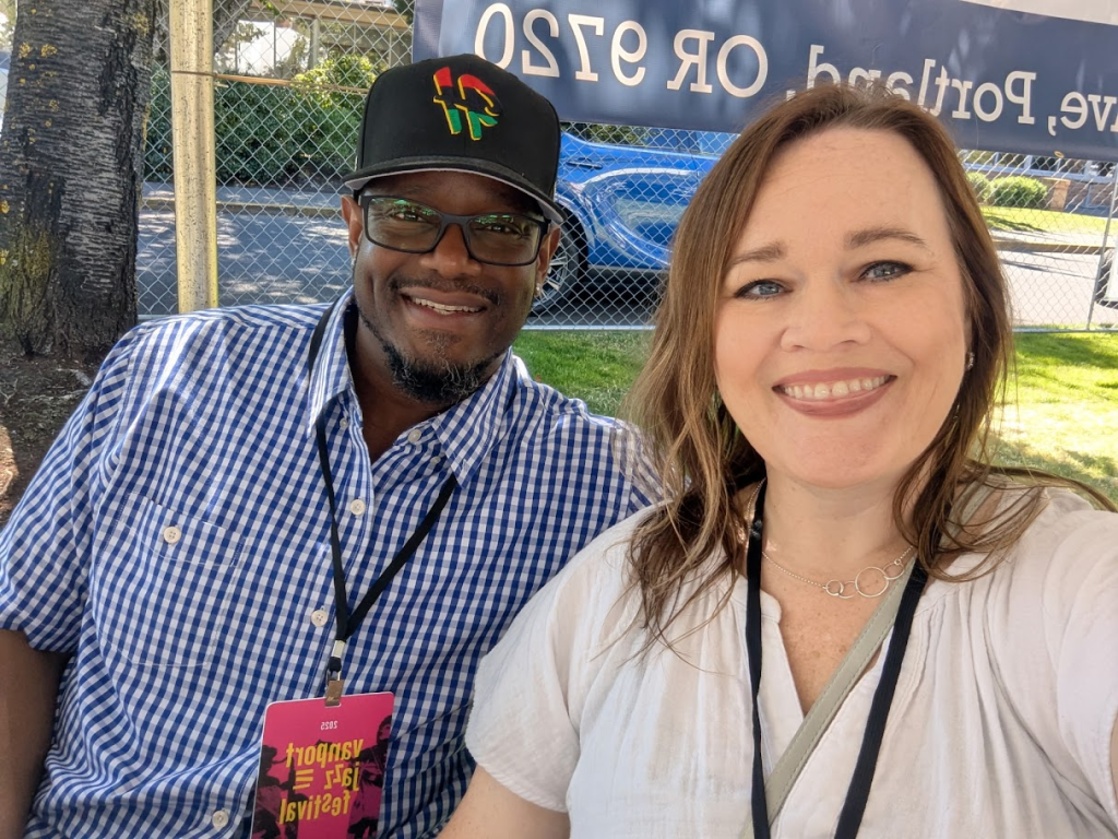 A man and a woman smile at the camera while sitting outdoors. The man wears glasses, a black cap, and a blue checkered shirt with a festival badge. The woman has long brown hair and wears a light-colored top. A fence is visible behind them.