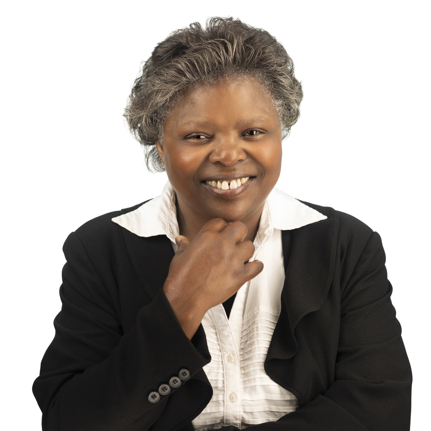 A smiling woman with short, graying hair wearing a black blazer over a white shirt, sitting and resting her chin on her hand against a plain white background.