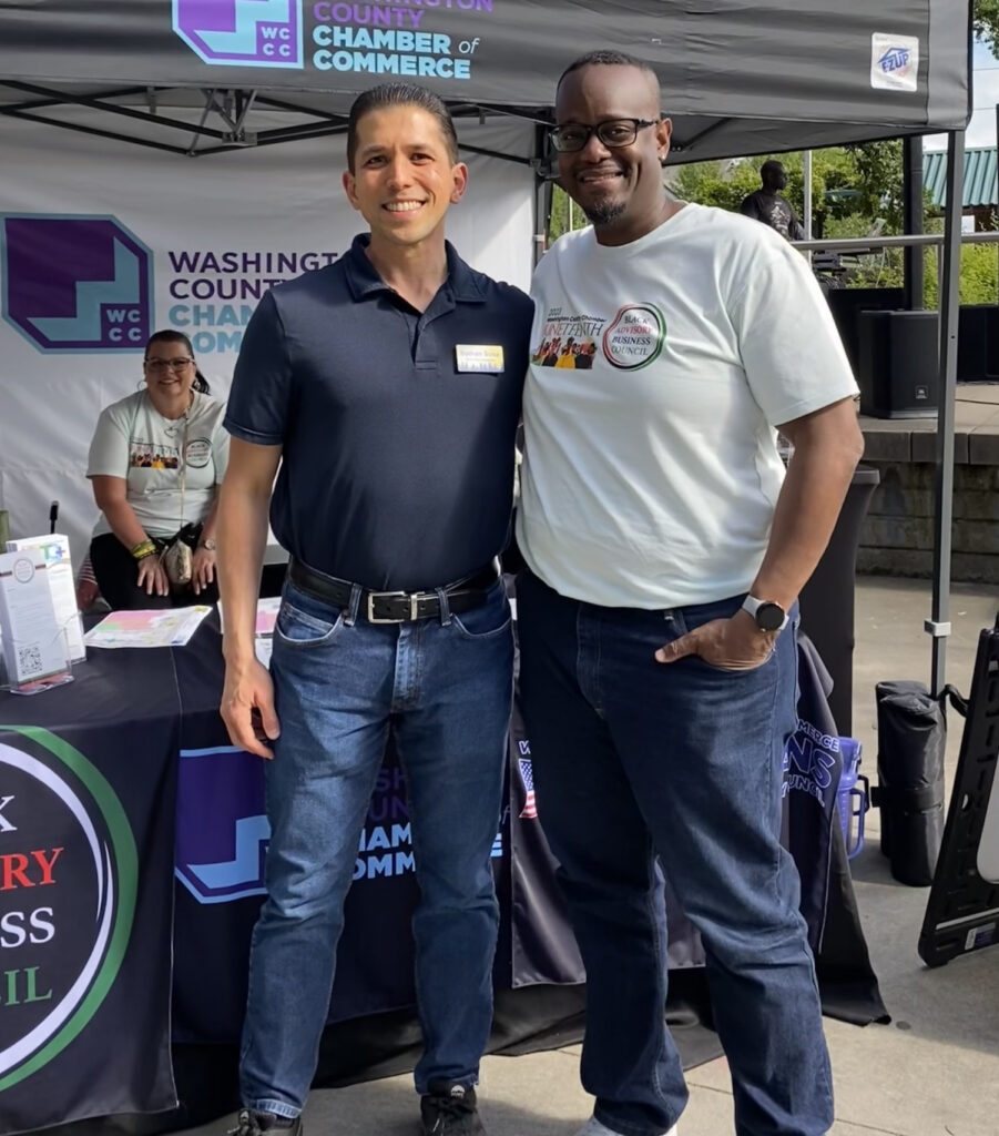 Two men smile and pose in front of a Washington County Chamber of Commerce booth at an outdoor event. A woman sits at the booth under a canopy, and trees and other event signs are visible in the background.