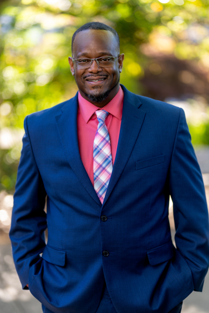A man wearing a blue suit, pink shirt, and plaid tie stands outdoors, smiling with his hands in his pockets. The background is blurred greenery and sunlight.
