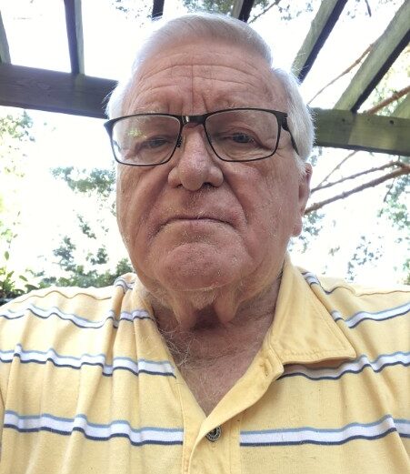 An older man with glasses wearing a yellow, striped polo shirt sits outdoors under a wooden pergola, with trees and sunlight in the background.