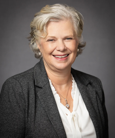 An older woman with short, wavy gray hair smiles while wearing a dark gray blazer over a white blouse. She has a necklace and is posed in front of a plain gray background.