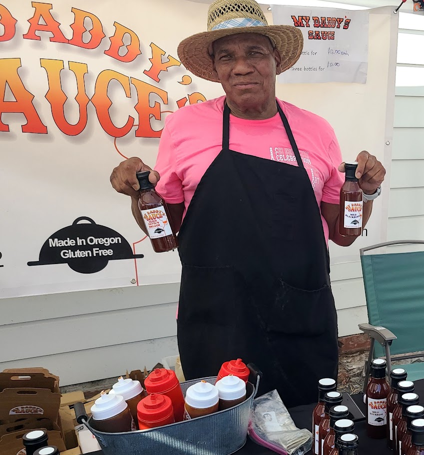 A man wearing a straw hat, pink shirt, and black apron stands at a booth holding two bottles of sauce. The booth displays a sign for "Daddy's Sauce" and several sauce bottles are arranged on the table.