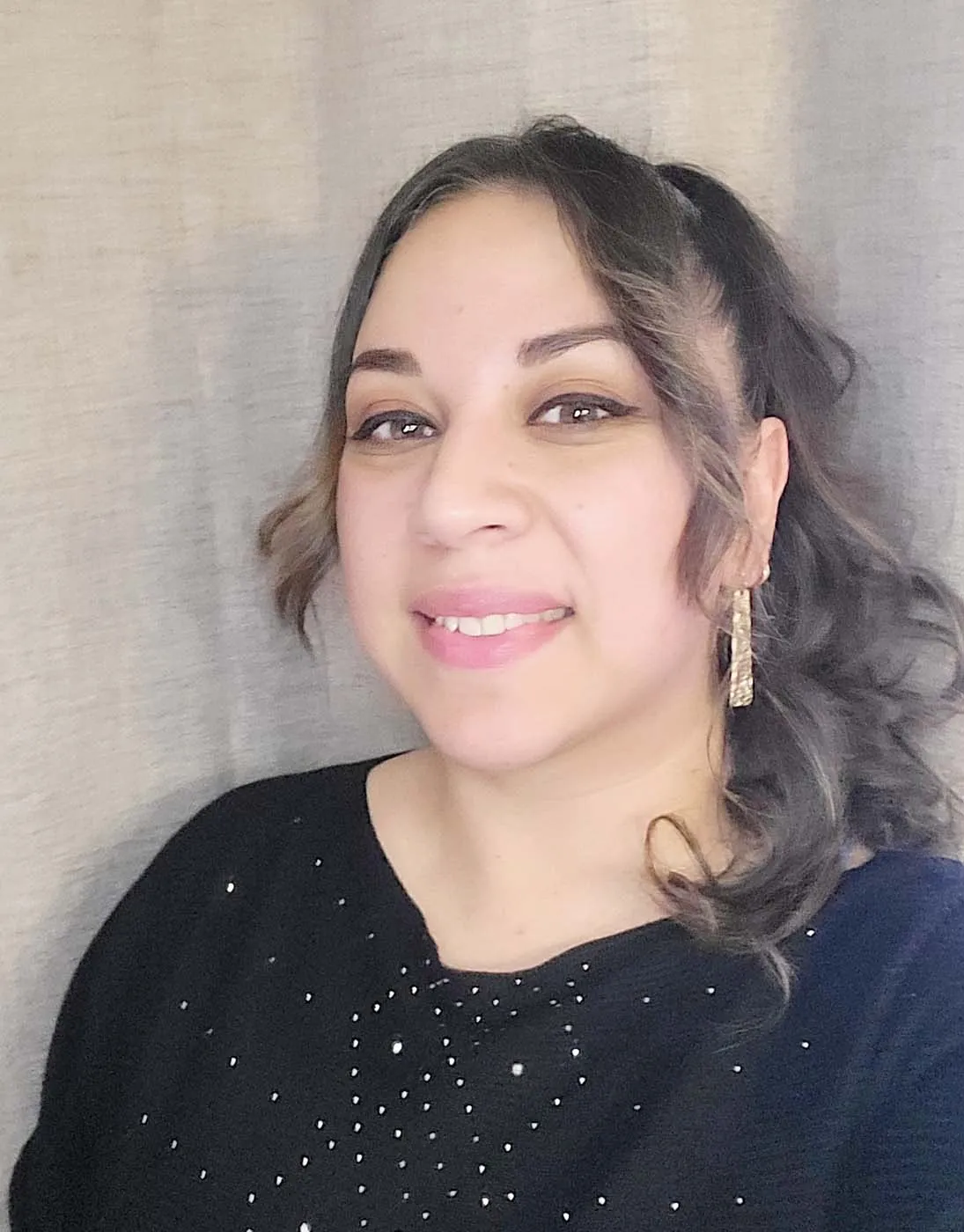 A woman with long, wavy hair pulled to one side, wearing gold earrings and a black top with small shiny embellishments, smiles in front of a neutral, light-colored background.