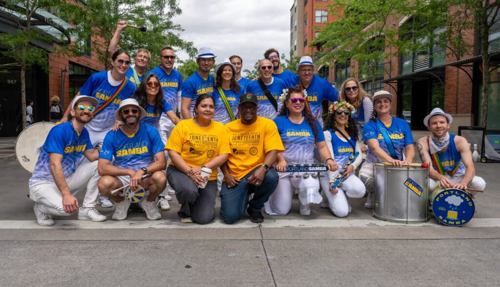 A group of people in blue, white, and yellow shirts pose on a city street with musical instruments, including drums, smiling and appearing festive outdoors with trees and buildings in the background.